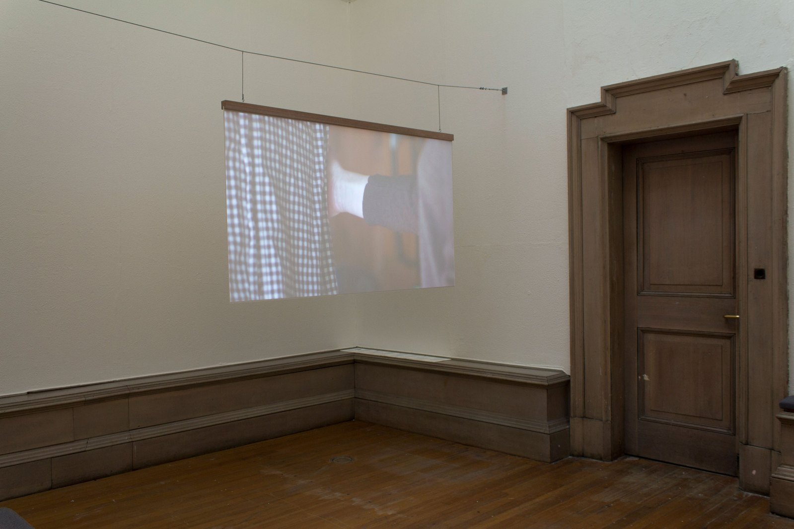 A hanging projection screen inside a beige room with a door to the right In the projection someones arm is reaching to the touch another persons arm who is wearing a checkered shirt