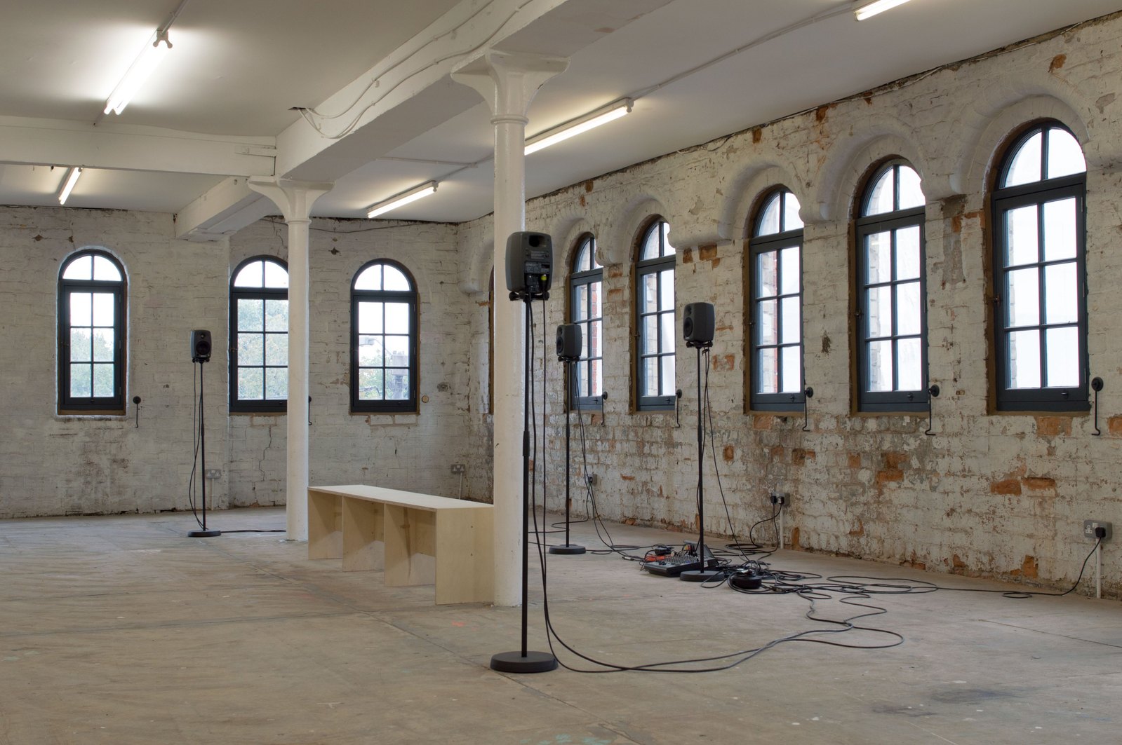 An old factory building with arches windows, white pillars and plywood floor. There is a wooden bench and an audio system. Jamie Kane