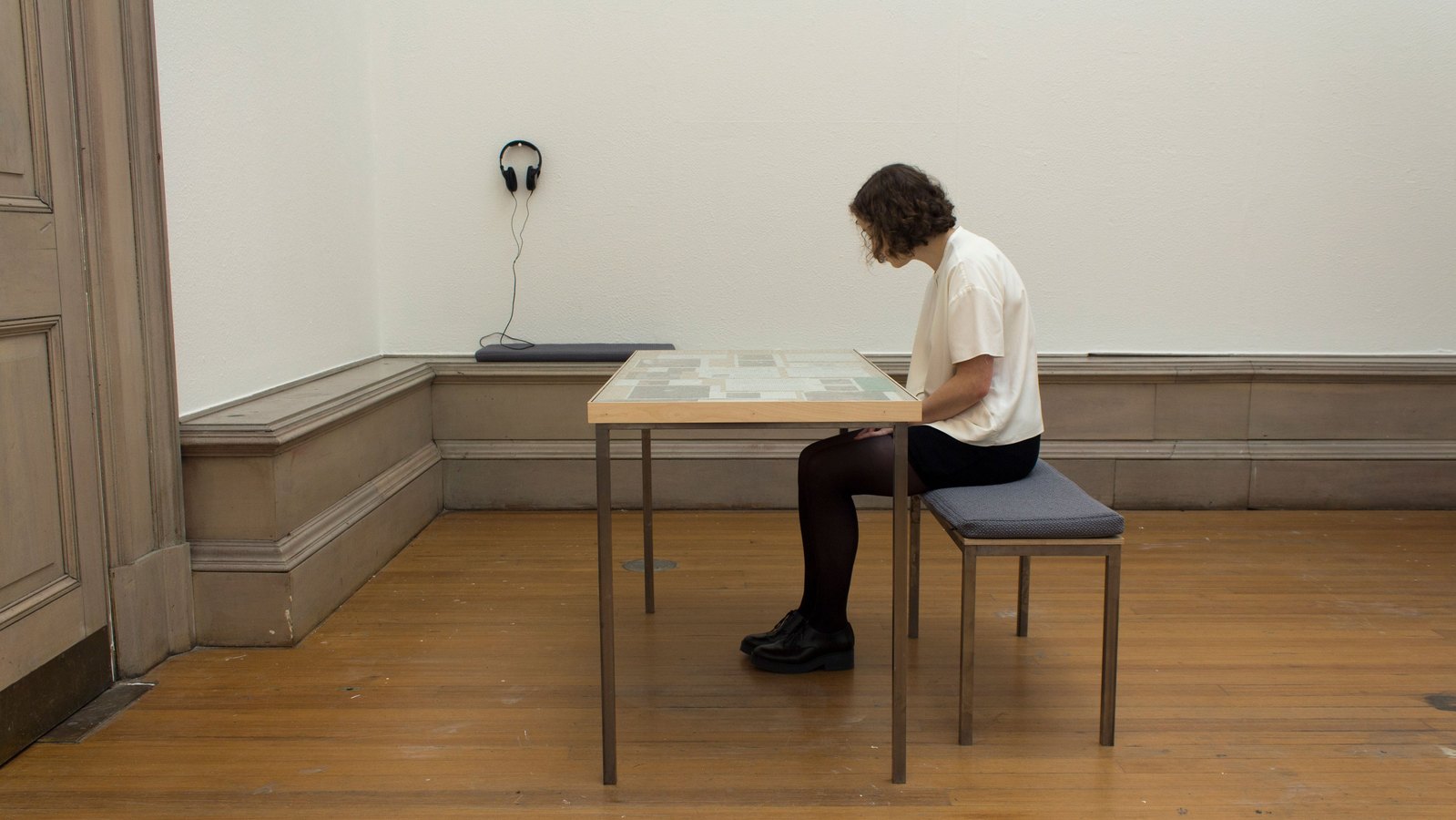 A beige room with wooden table. A woman with blonde hair, wearing white is sitting on a blue bench looking at the table