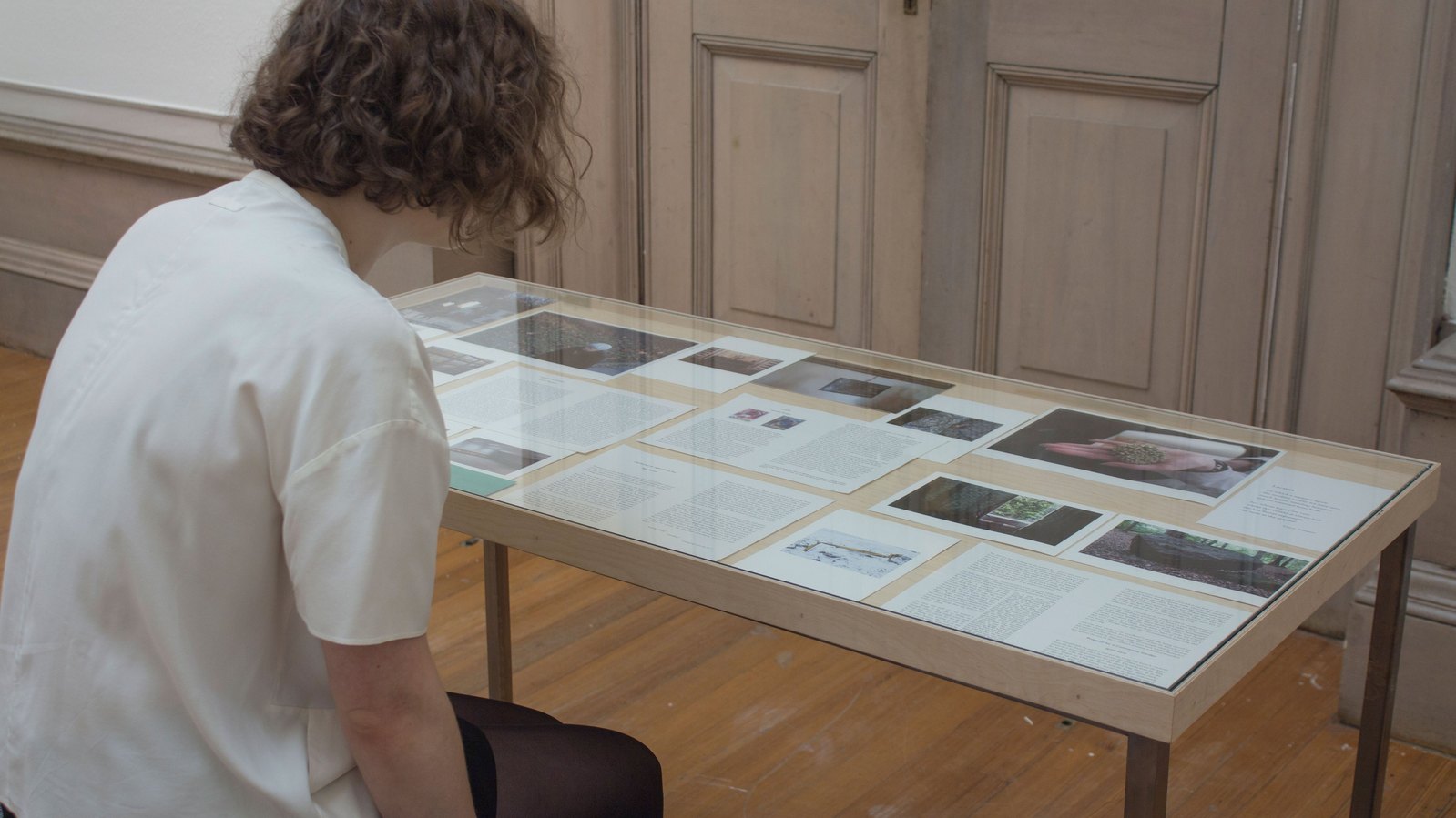 A wooden table with sheets of paper containing images and writing under glass. A woman with blonde hair, wearing white faces away toward the table whilst sitting