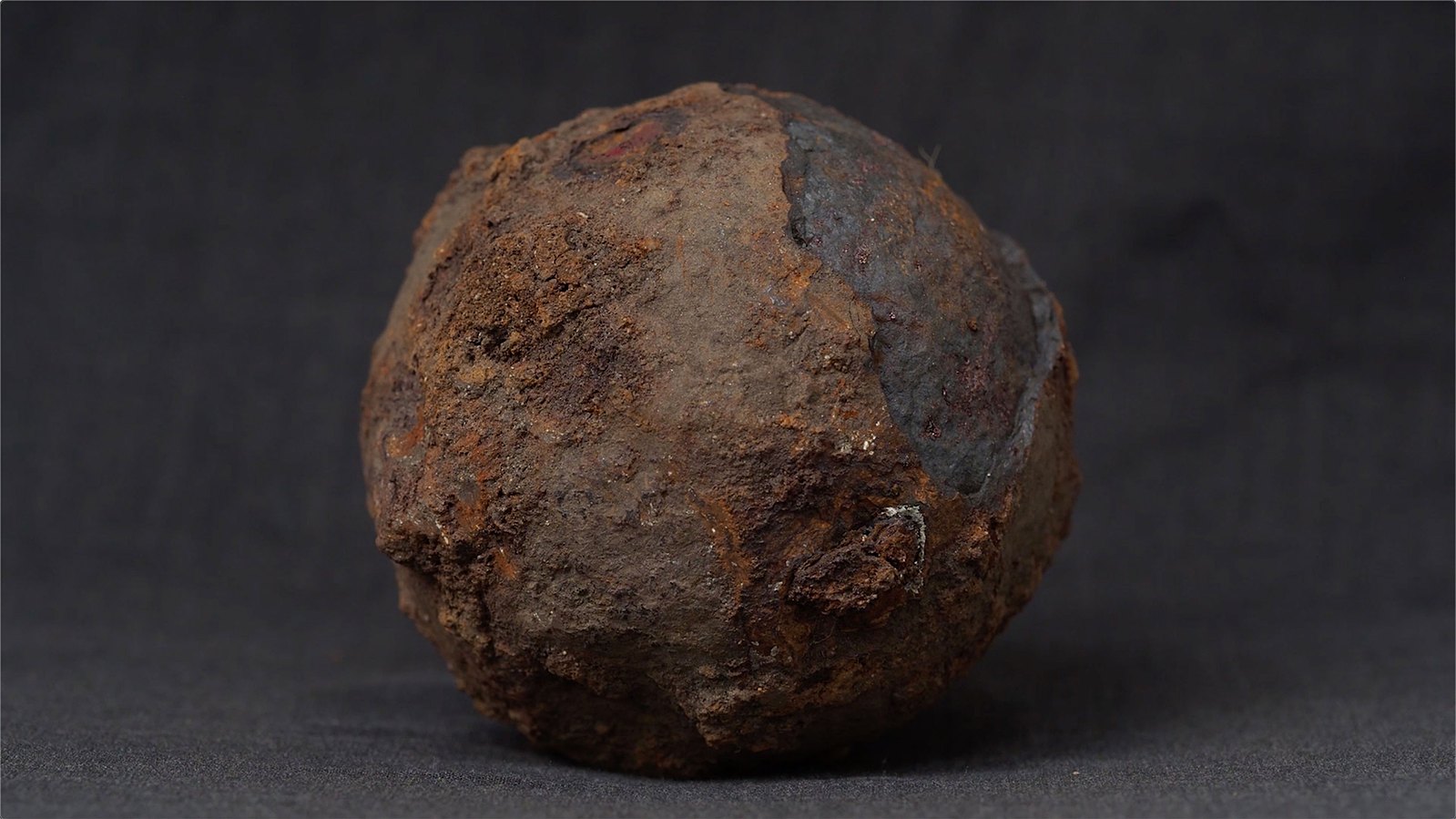 A rusted iron cannonball, with a dark grey fabric background. Jamie Kane