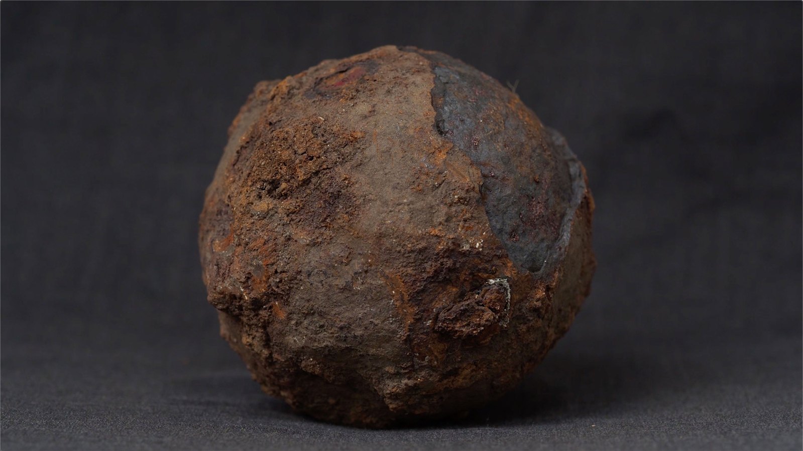 A rusted iron cannonball, with a dark grey fabric background. Jamie Kane