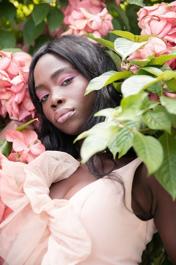 Dark skin model @Taylume wearing pale blush coloured Love and Lemons dress surrounded by foliage and pink and peach flowers, photographed by Sharleen Christie Photography.