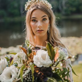 Natural bridal makeup and hair model wearing glass bead headband and holding large white and green bouquet in a country setting.
