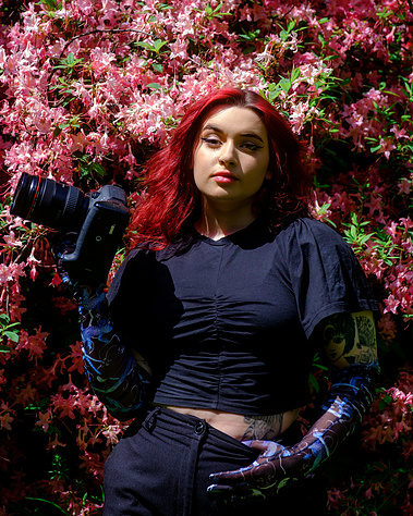Portrait of a young, women with striking eyeliner, bright red hair and posing with a large Canon camera in front of a pink floral wall. This portrait is the owner of Carla Marcia Photography. 