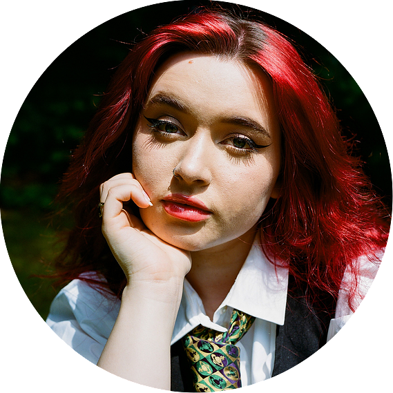 Portrait of a women wearing a white shirt and green monogram tie with a black waistcoat. She poses calmy with her hand against her cheek and she has bright, fiery cherry red hair. There is a slight grain to the image.
