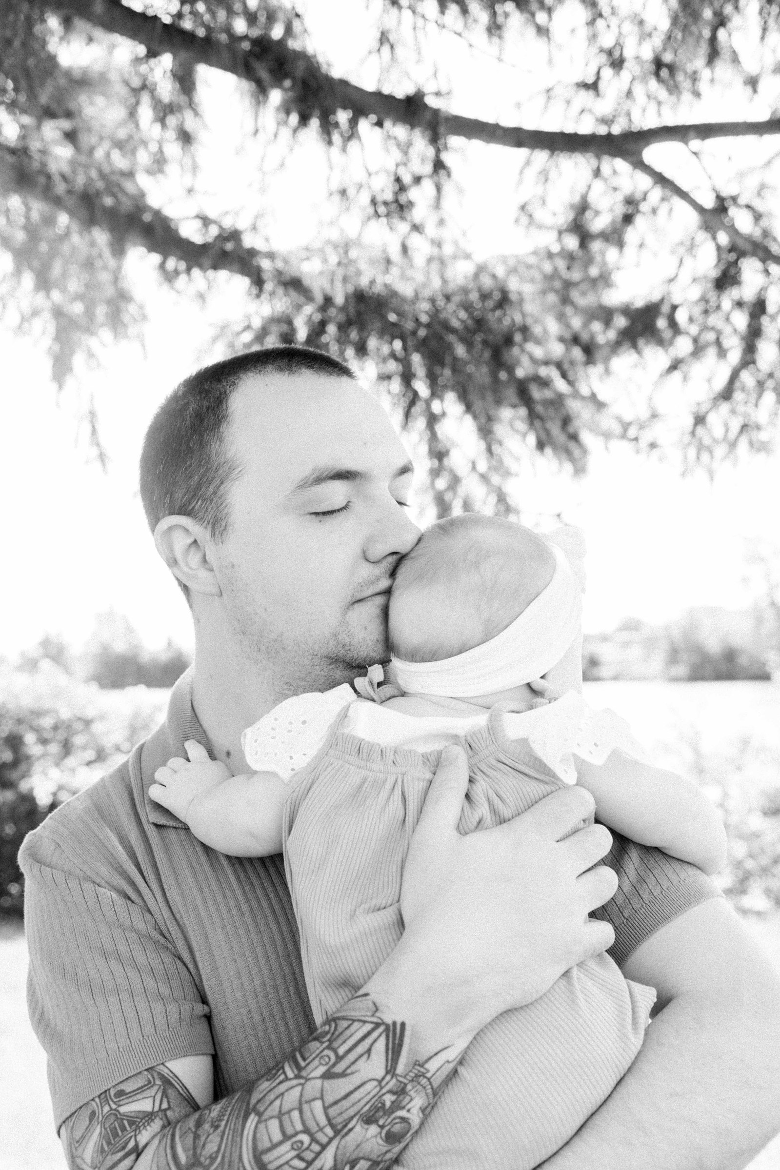 Black and white candid portrait of a dad kissing his baby daughter in front of the Ottawa River in Hawkesbury, Portrait Photographer, Family Photographer, Emily VanderBeek Photography, Candid Photography, making memories.
