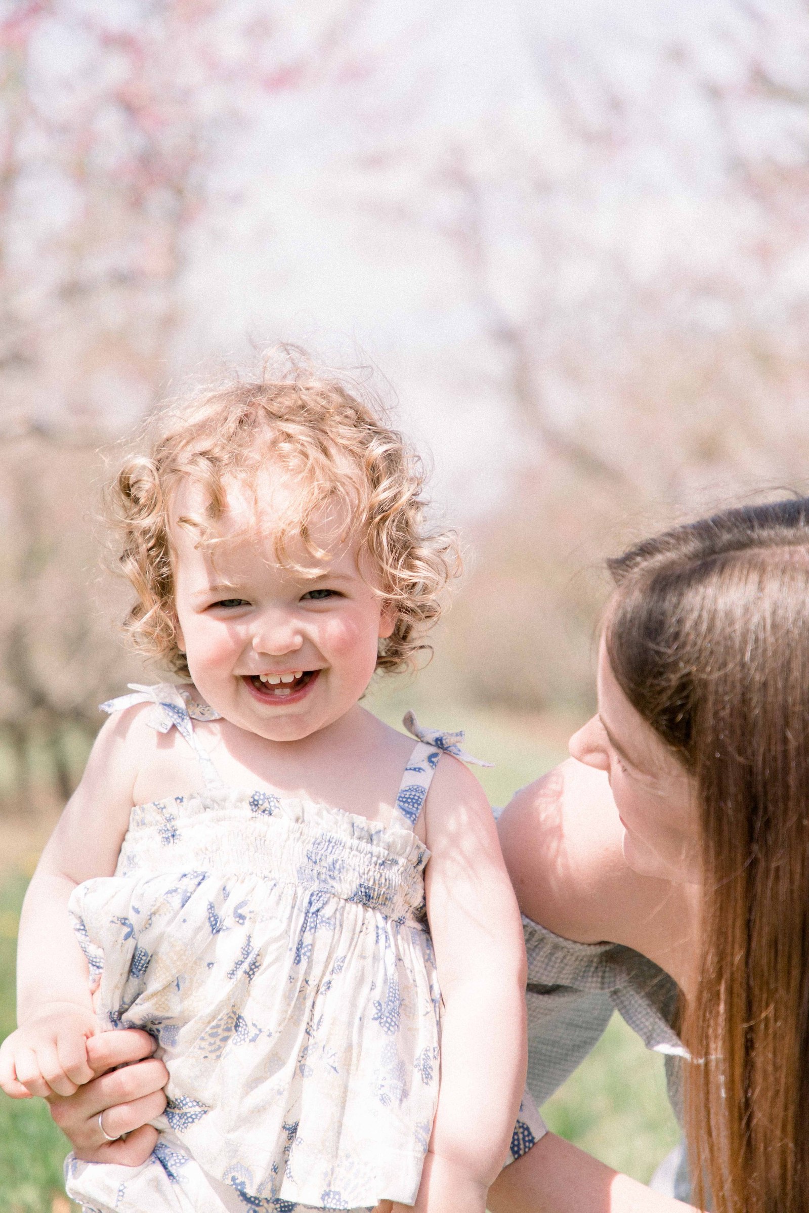 Portrait of mother and baby daughter hugging and laughing in an orchard of fruit blossoms. Niagara Family Photographer, Niagara Portrait Photographer, Niagara Motherhood Photographer. Emily VanderBeek Photography, Emily VanderBeek Photo.