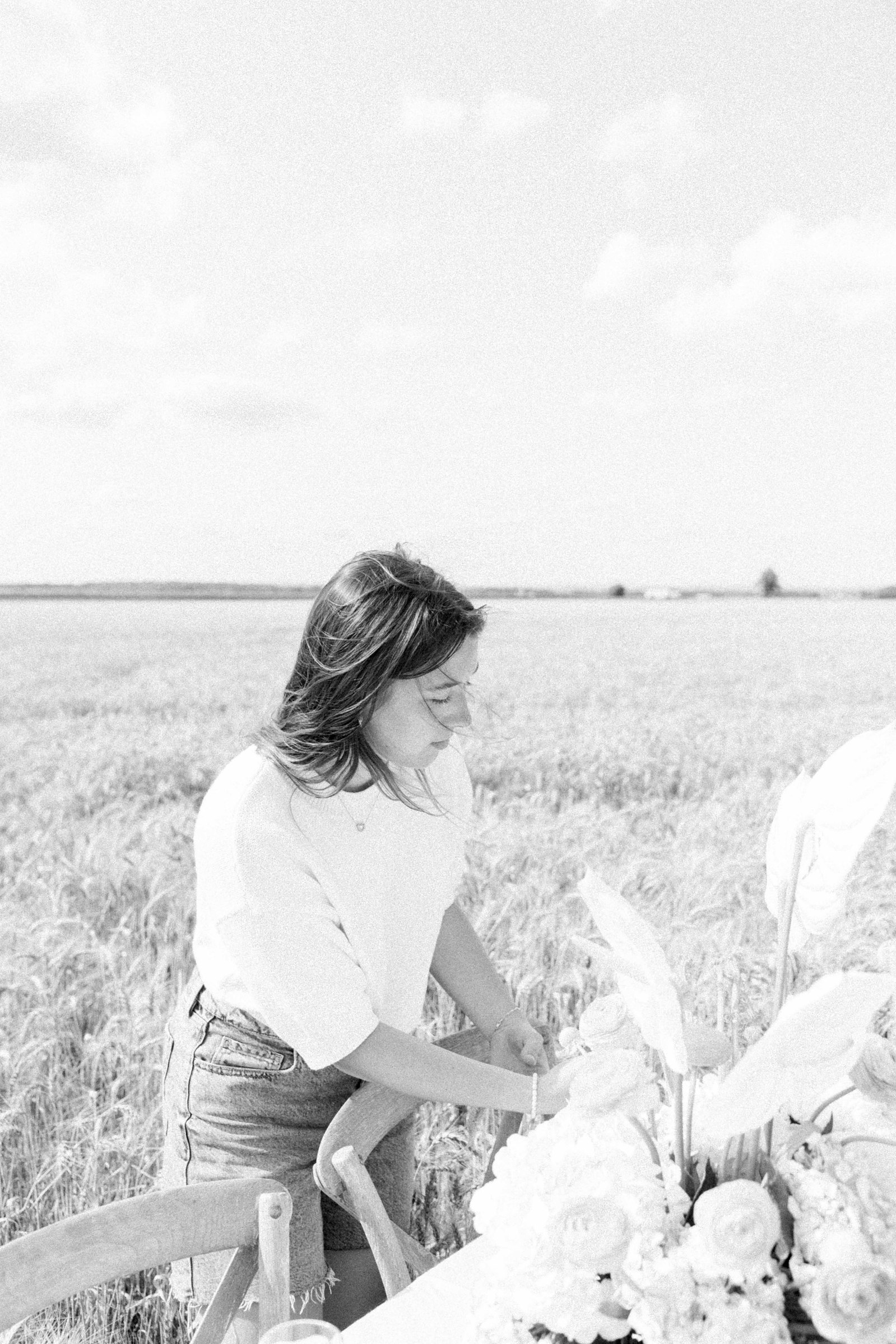Black & white photo of a woman working on a table setting in a wheat field in Rigaud, Quebec. Emily VanderBeek Photography. Montreal Photographer. Light & Airy photographer.