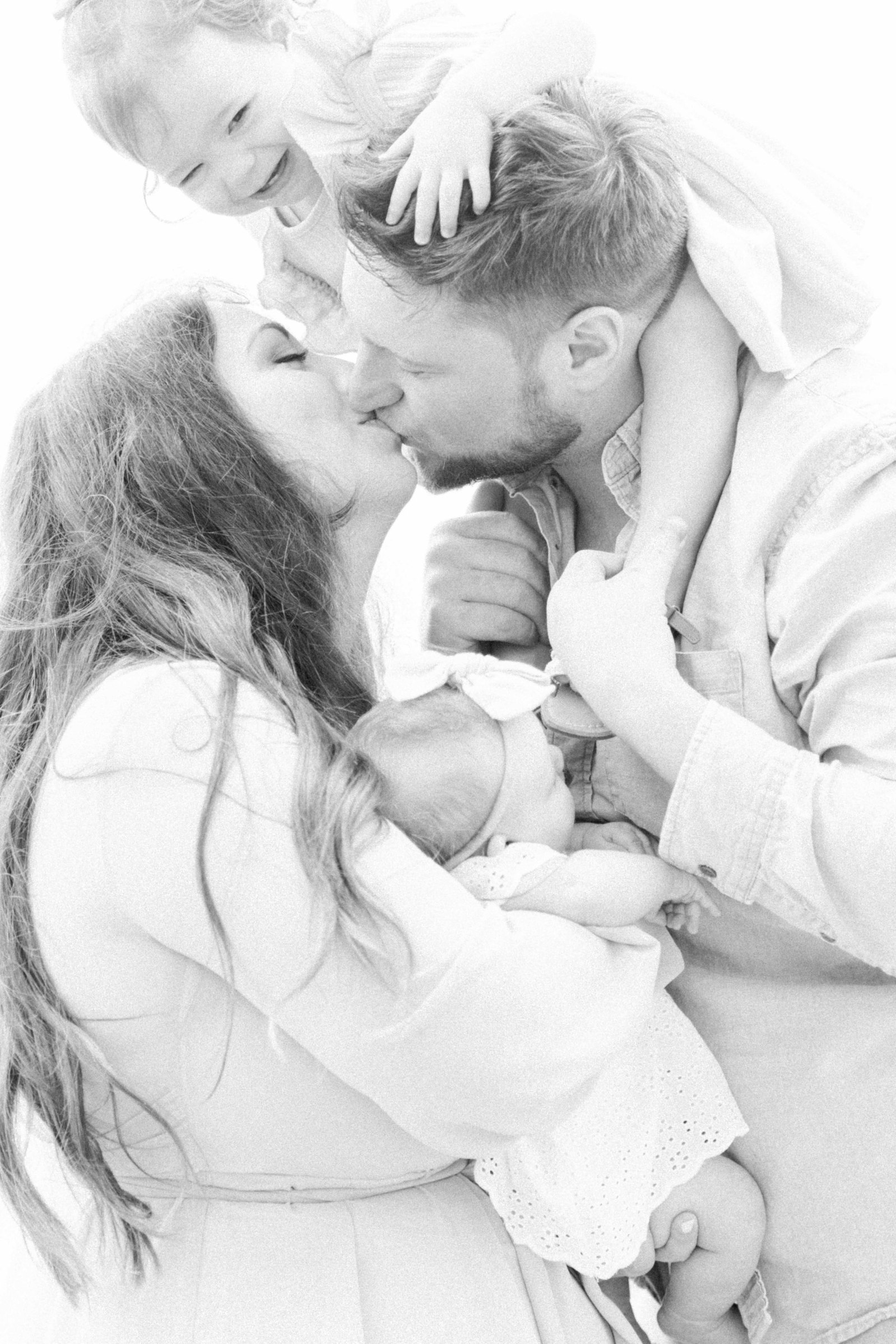 Black and white portrait of family of four on the beach, husband and wife kissing, Niagara Portrait Photographer, Niagara Family Photographer, Niagara Motherhood Photography, Beach Portrait Photography, Emily VanderBeek Photography.