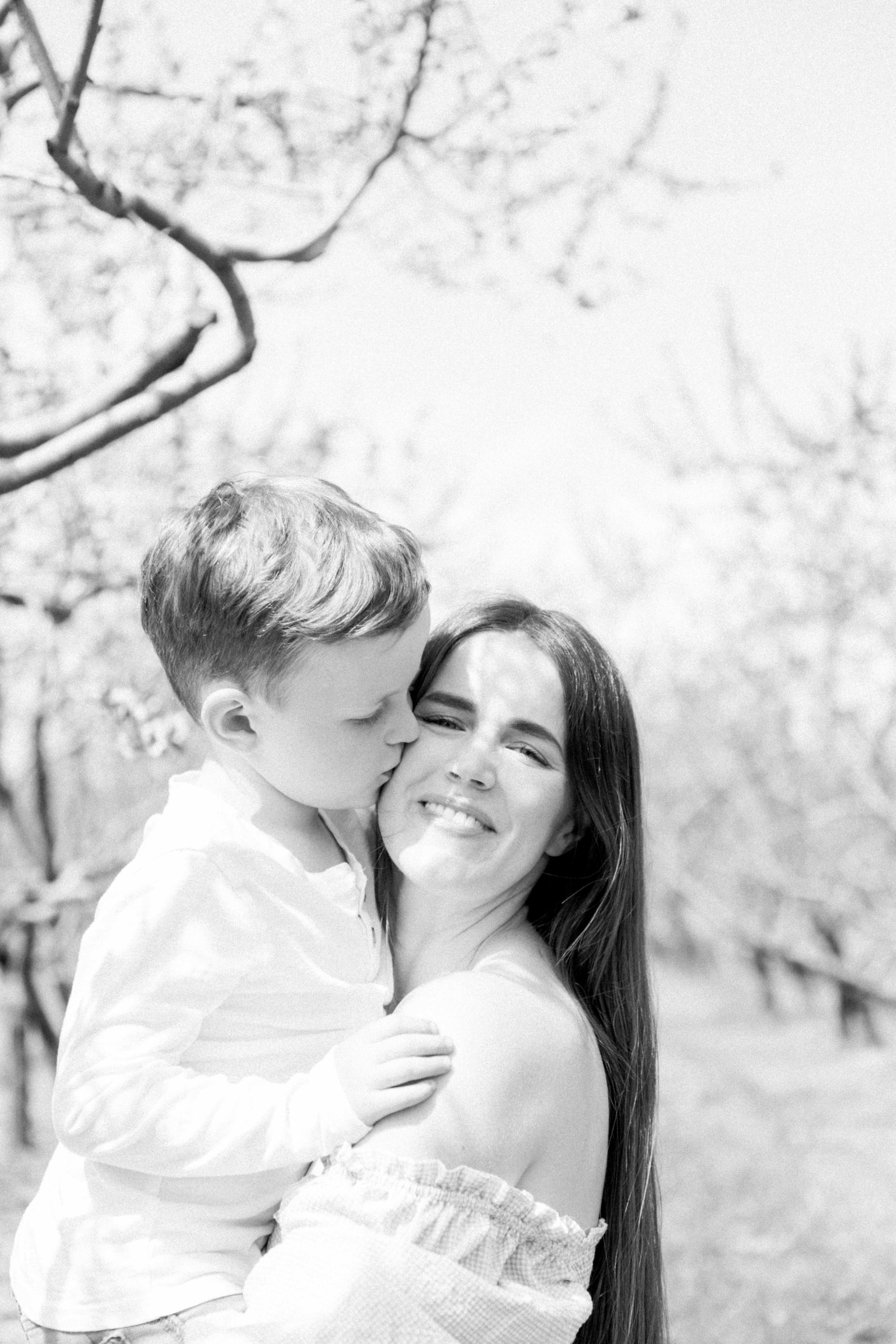 Black and white portrait of mother and son hugging in an orchard of fruit blossoms. Niagara Family Photographer, Niagara Portrait Photographer, Niagara Motherhood Photographer. Emily VanderBeek Photography, Emily VanderBeek Photo.