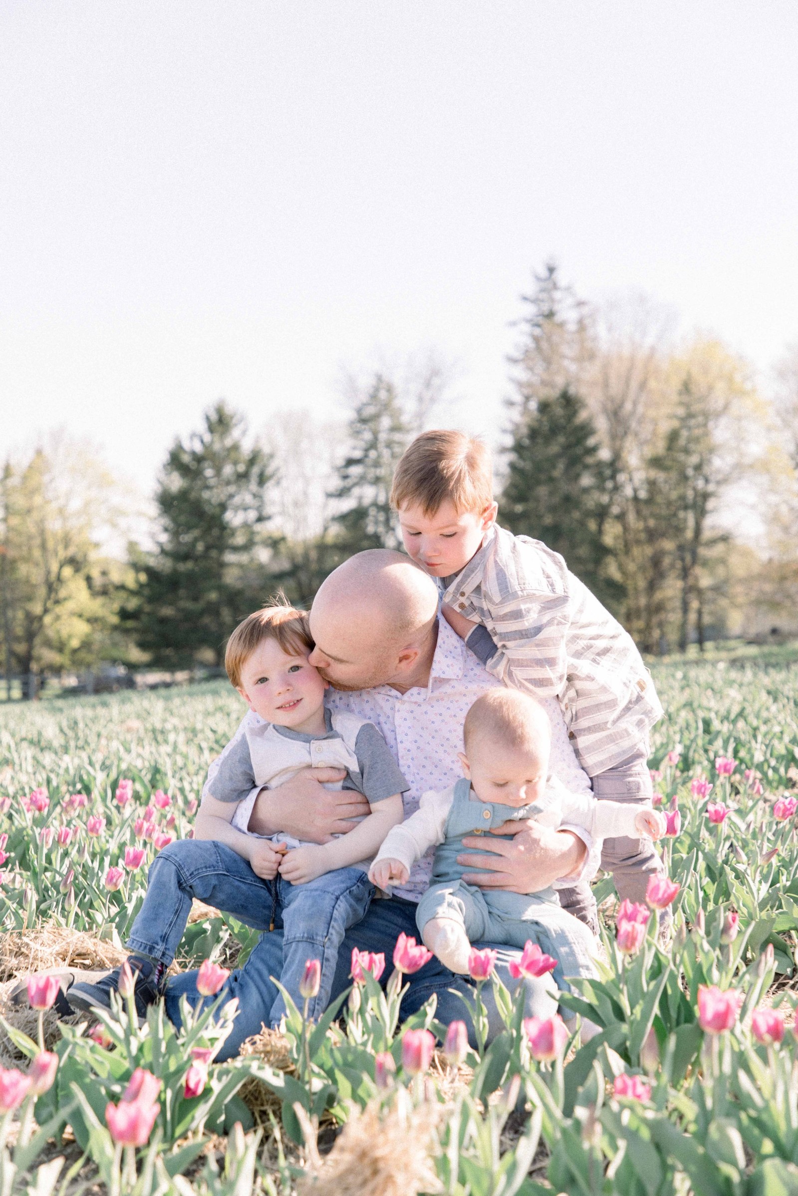 Portrait of father with three sons sitting in a tulip field. Father is kissing one son on the cheek. Vankleek Hill Portrait Photographer, L'Orignal, Champlain, Prescott-Russell, Family Photography, Candid Photography, Emily VanderBeek Photography.