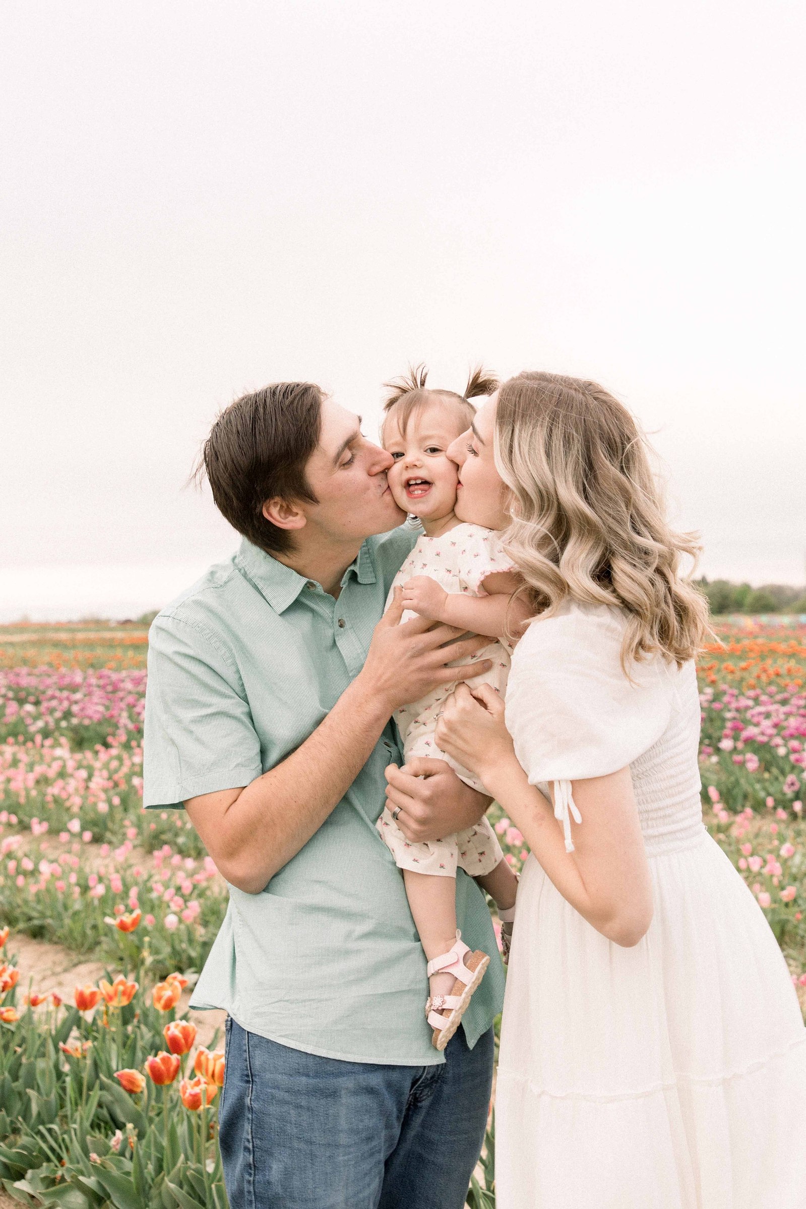 Portrait of a family of three at a tulip field in Niagara, kissing their daughter on the cheeks. Niagara portrait photographer. Niagara family photographer. Niagara photographer. Emily VanderBeek Photography. Candid photography. Light and airy photography