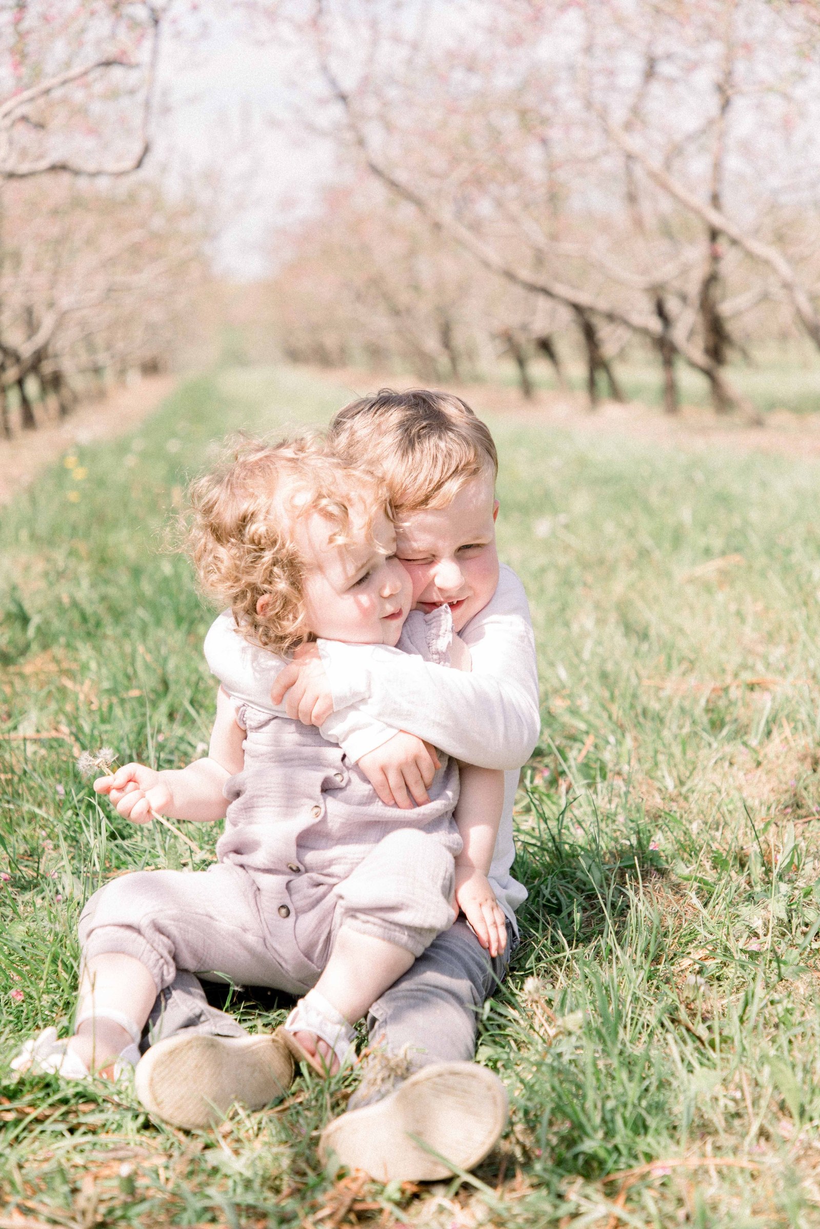 Portrait of brother and sister hugging and smiling in an orchard of fruit blossoms. Niagara Family Photographer, Niagara Portrait Photographer, Niagara Motherhood Photographer. Emily VanderBeek Photography, Emily VanderBeek Photo.