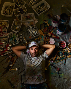 Environmental portrait of mural artist Rafael Blanco lying on a bed of paint brushes surrounded by paint trays and art supplies. 