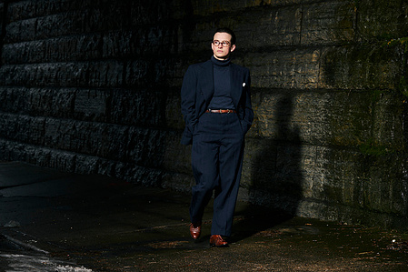 environmental fashion portrait of Zoë Vulgamott walking on a side walk under a bridge wearing a dark navy blue suit and brown dress shoes. 