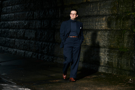 environmental fashion portrait of Zoë Vulgamott walking on a side walk under a bridge wearing a dark navy blue suit and brown dress shoes.