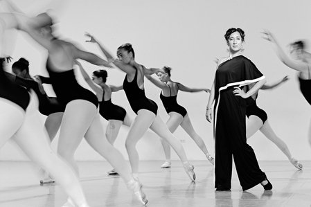 Black and white environmental portrait of a female ballet dance director surrounded by ballet dancers, blurred with slow shutter speed. Photographed in Chicago for Chicago Magazine