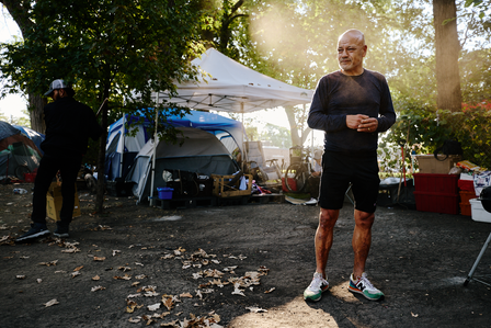 Environmental portrait of a homeless, houseless man, in an encampment, in front of his tent. Sun shining from behind him silhouetting him. 