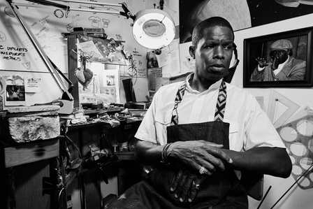 Black and white environmental portrait of a man in his small jewelry workshop in a room in his home. Photographed in Buffalo New York
