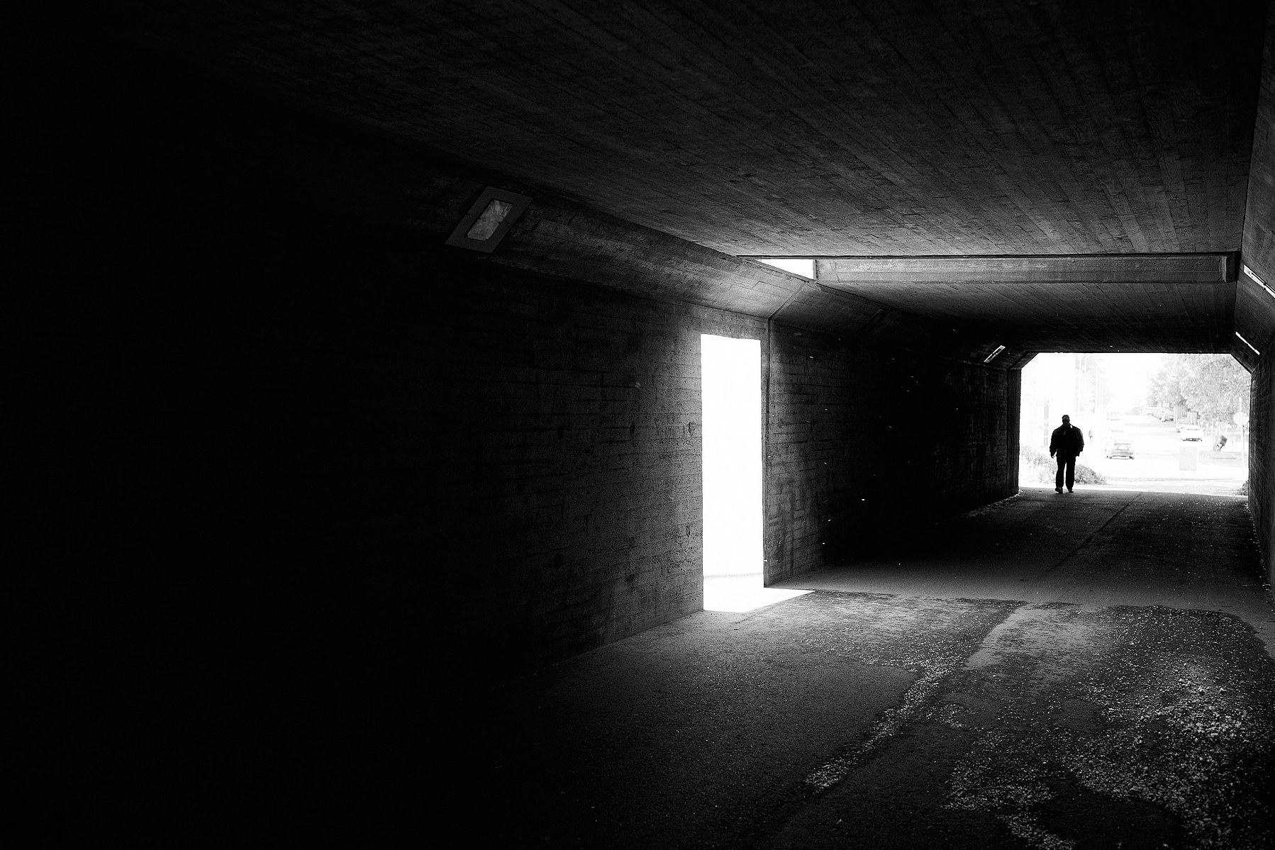 Man walks through dark underpass, in the foreground sun light is reflecting from the dark wall, resembling door