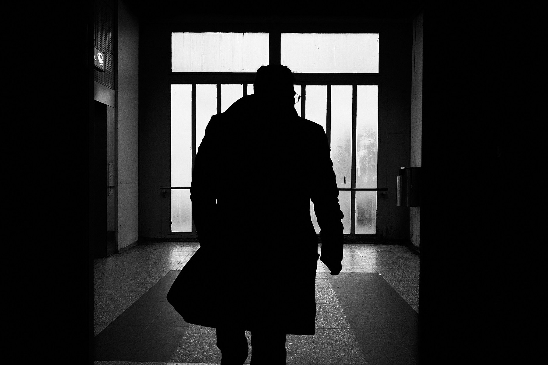 A man wearing trench coat walks at Malmi train station, large window is behind the man