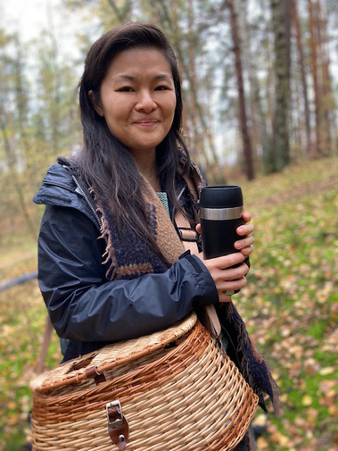 A friendly woman with a warm smile standing amidst the forest, carrying a mushroom basket. She is your knowledgeable guide, ready to lead you on an exciting mushroom adventure.