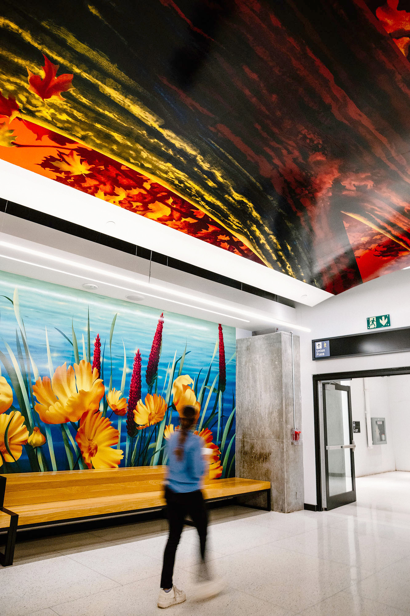 A woman walks across colourful murals and artworks depicting nature and plants are displayed in a public setting.