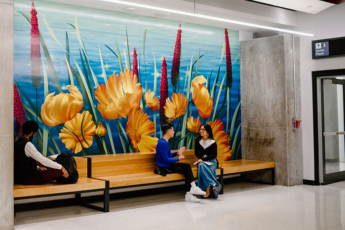 Two people converse behind a bench adorned with a vibrant mural of flowers and plants in a public space.