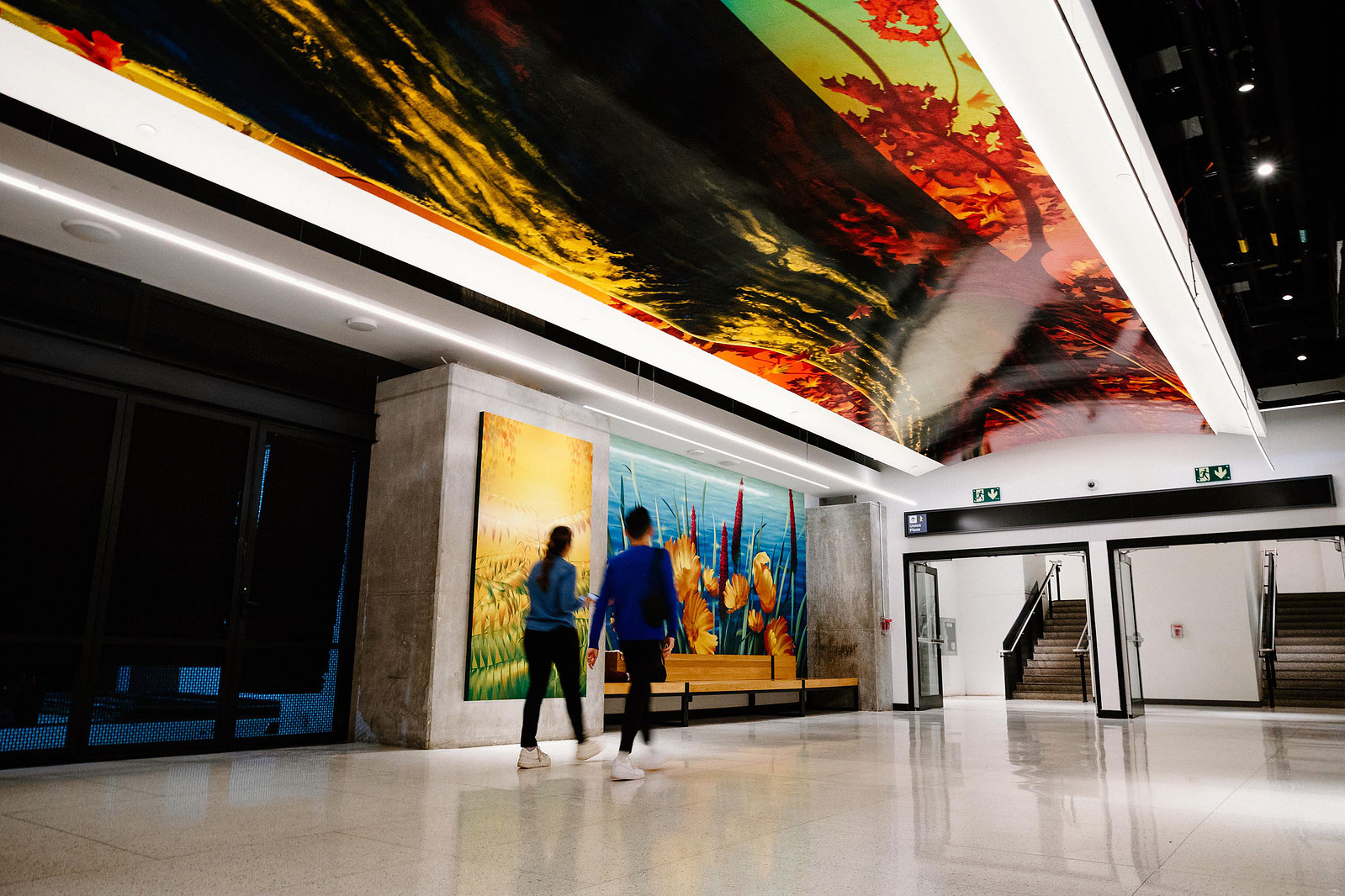 Two people walk across colourful murals and artworks depicting nature and plants in a public setting.