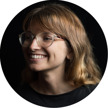 A photo of Alex dressed in a black shirt against a black background. She is looking to the left and smiling, she is white, has long brown hair and is wearing clear frame glasses.