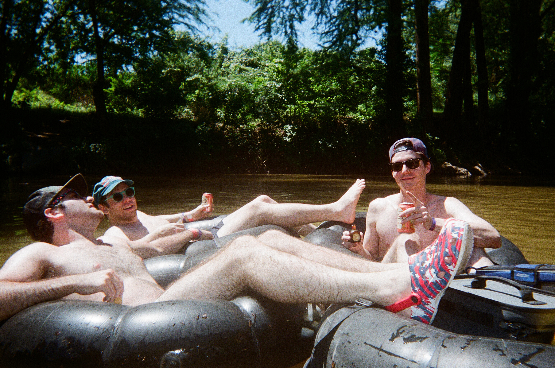 Three friends in inner tubes, floating the Comal River. These guys' have WHITE legs, and one's Americana Croc-like shoes really stand out in this photo. 