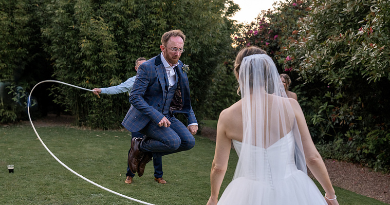 Groom jumping a skipping rope.  His wife and a best friend are twirling the rope.