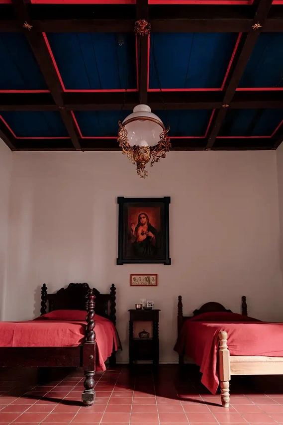 In one of the guest rooms, a reproduction of a Sacred Heart of Jesus painting and an Art Nouveau glass and brass lighting fixture over twin beds; the night stand and the bed on the left are made by the Mexican artist León Muñiz. 
