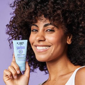 Curly haired Model smiling and holding Noughty haircare, The Saviour universal balm.
Photographed and styled by Tom Hargreaves in a studio in London.