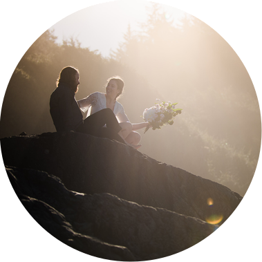 An eloping bride and groom sit together atop a large rock on a ruggedly beautiful Northern California beach
