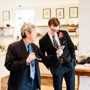 A groom and his father talk before the wedding ceremony; the father shows the groom something on his phone