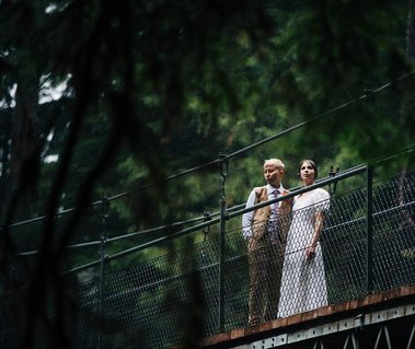 An eloping couple stands together on high bridge, looking out at the forest around them
