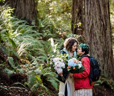 An eloping couple stands on a hiking trail, wearing warm, rugged coats over their bridal gowns