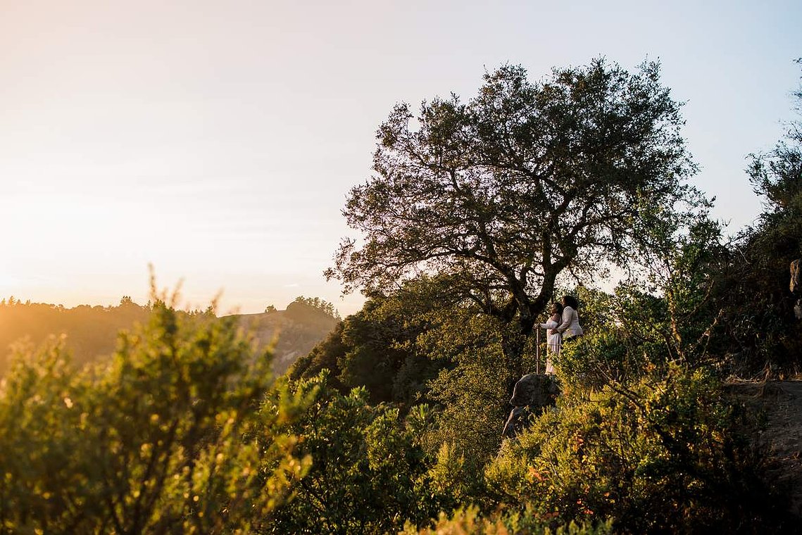 A pair of brides stands at a rocky viewpoint, gazing out at the sunset and the mountains around them