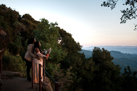 Two brides gaze out at a mountainous, forested landscape from a rocky viewpoint