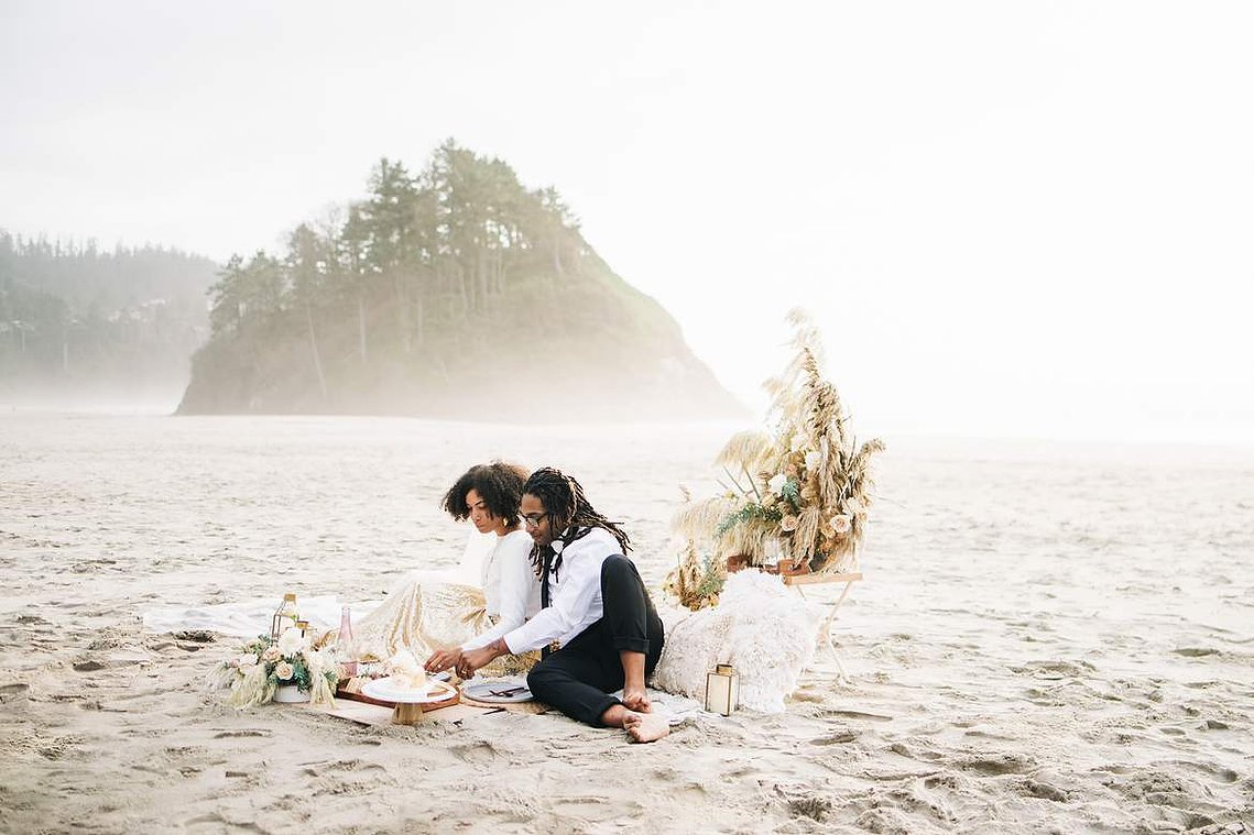 A bride and groom sit on a beach with an elaborately designed picnic; the groom cuts the couple's small but elegant elopement cake