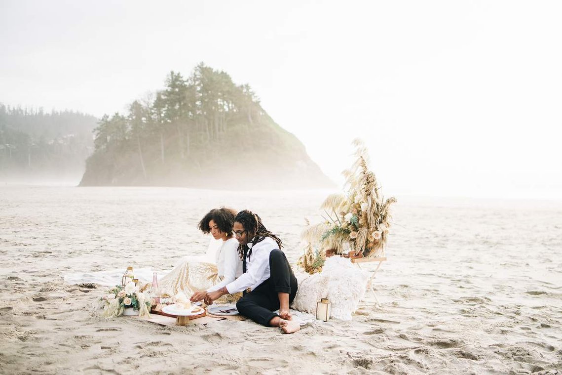 A bride and groom sit on a beach with an elaborately designed picnic; the groom cuts the couple's small but elegant elopement cake