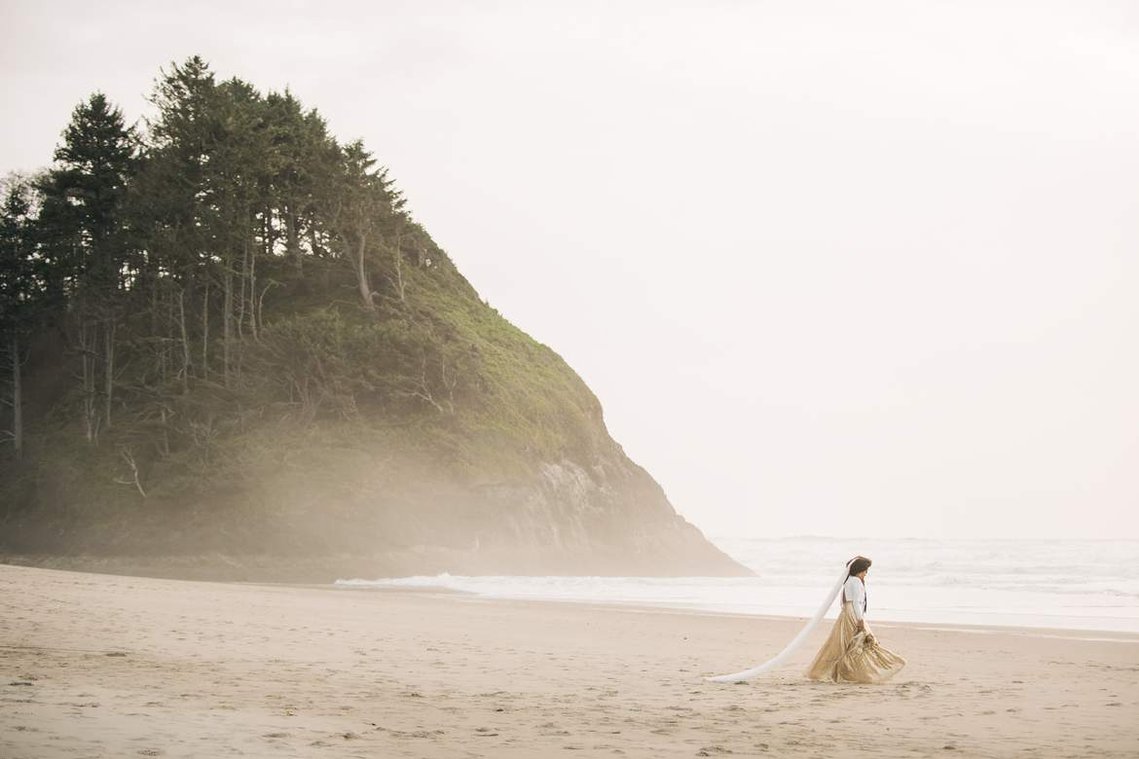 The bride and groom walk along the beach; we can see the mist coming in from the ocean and the bride's long veil trailing behind her