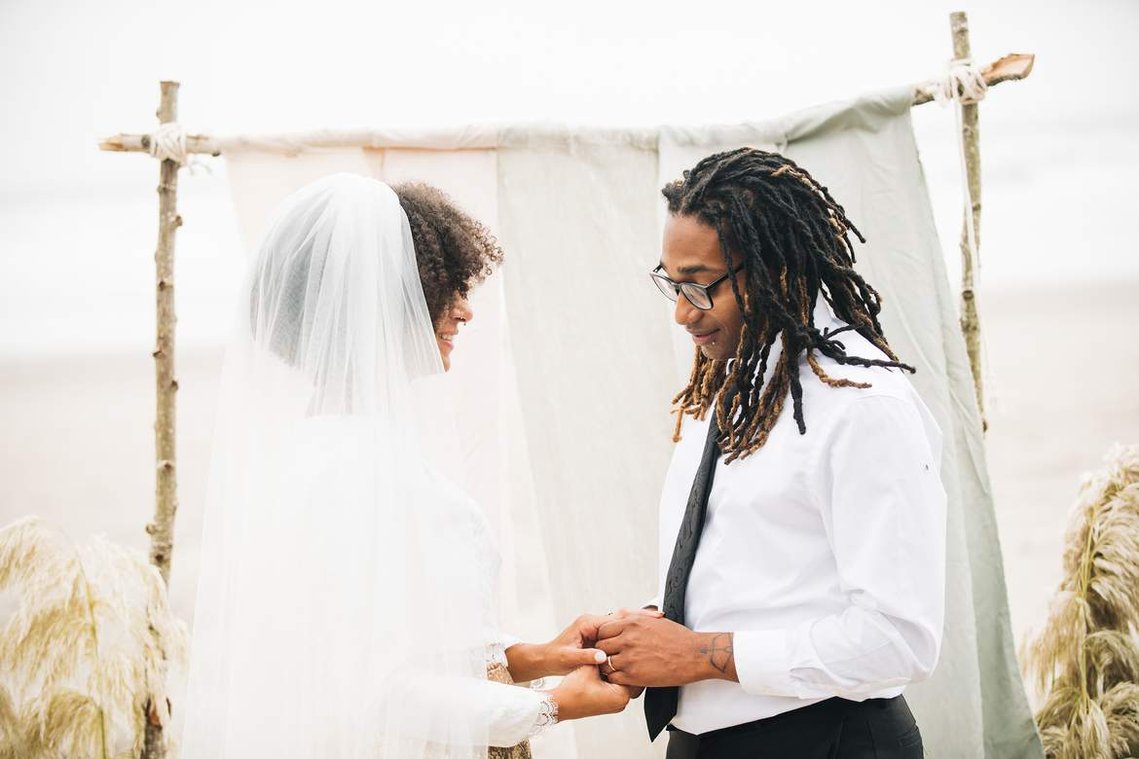During their elopement ceremony, the bride and groom stand in front of a driftwood arch; they clasp hands and look at each other