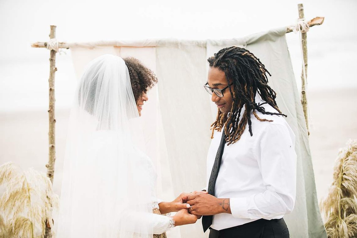 During their elopement ceremony, the bride and groom stand in front of a driftwood arch; they clasp hands and look at each other