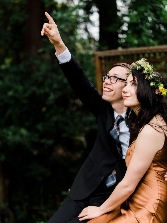 A bride and groom sit in their backyard after an intimate wedding ceremony at home; they look up at the trees around them and point