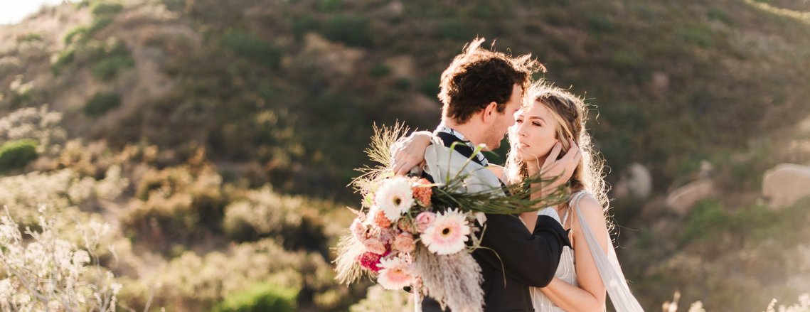 5 Secrets to Planning an Awesome California Elopement: A bride and groom stand together on a golden California hillside; the groom places a hand against the bride's face, tucking her hair behind her ear