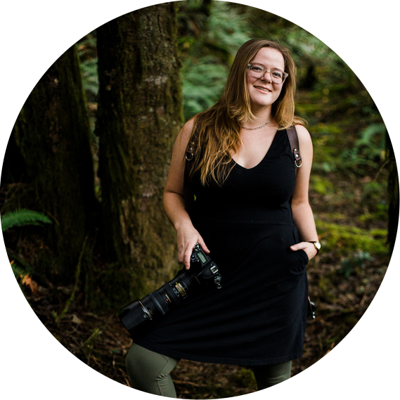 Portrait of Kate Waterman Rose: she stands in a deep green forest, holding two cameras and smiling at the viewer