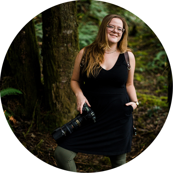 A portrait of the photographer, Kate Waterman Rose; she stands in a forest, carrying two cameras and smiling